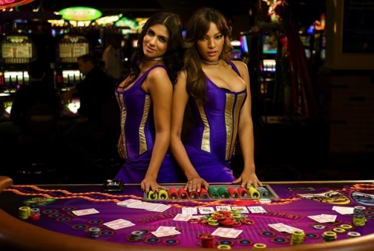 Две сексуальные девушки крупье позируют у стола для игры в блэкджек в фиолетовых обтягивающих мини-платьях